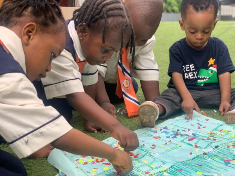 "Preschool students at Davebrook Schools sitting on green grass, working together to stick colourful paper bits onto a large house drawing during an art class with Ms. Abimbola."